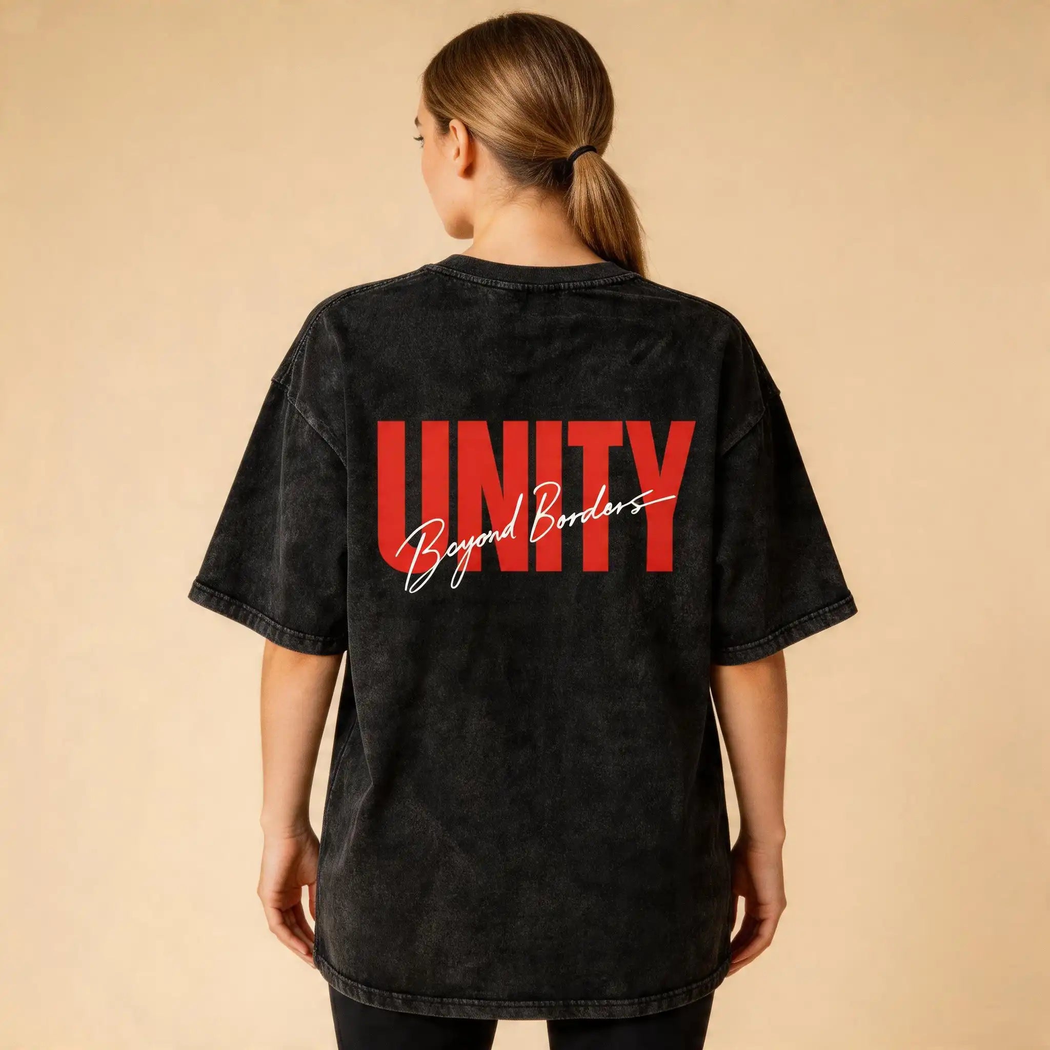 Back view of a female model wearing the Celvian Beyond Borders T-Shirt in washed black, featuring the bold red UNITY graphic with white Beyond Borders script on a clean beige studio background.