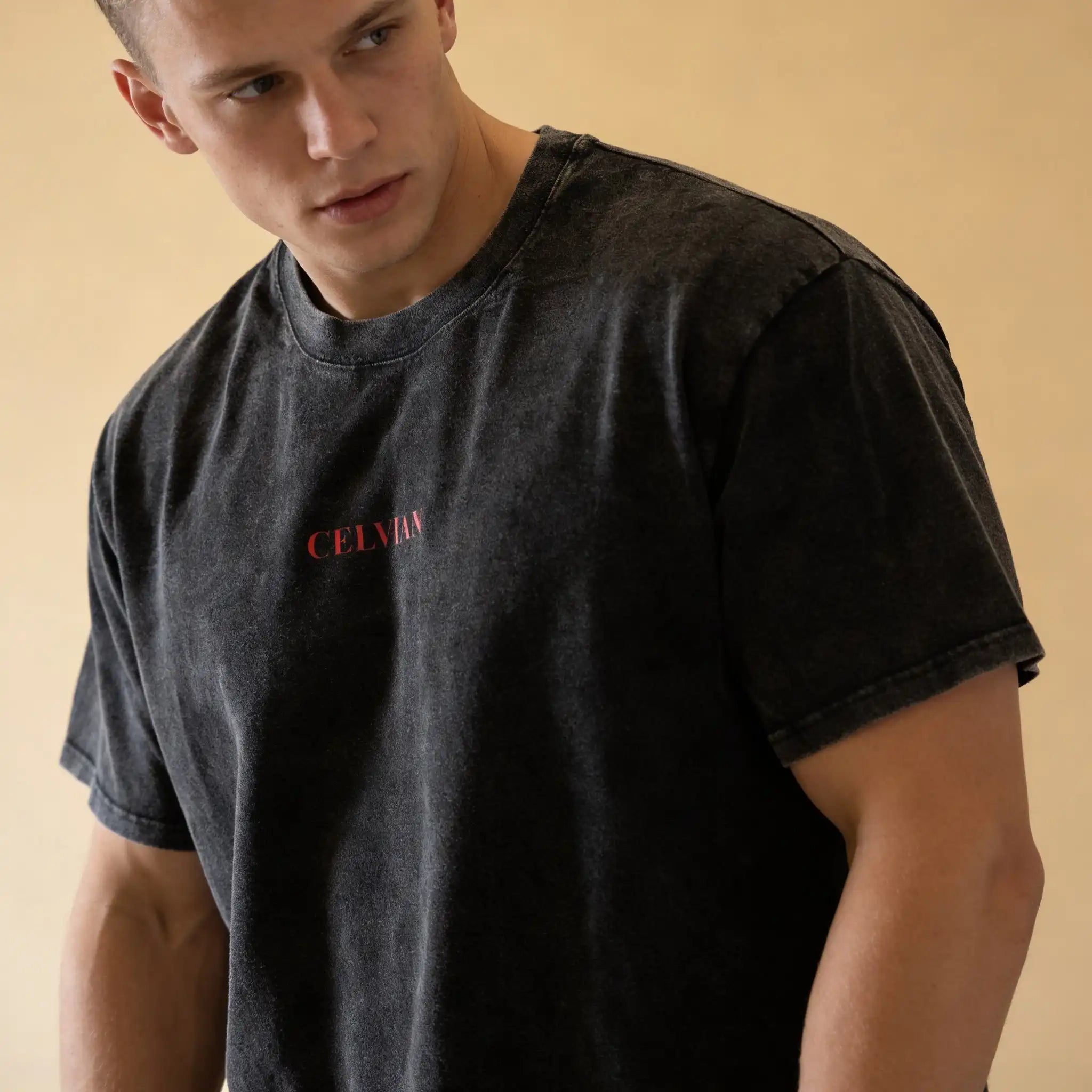 Close-up front view of a male model wearing the Celvian Beyond Borders T-Shirt in washed black, featuring a minimal red CELVIAN chest logo on a clean beige studio background.