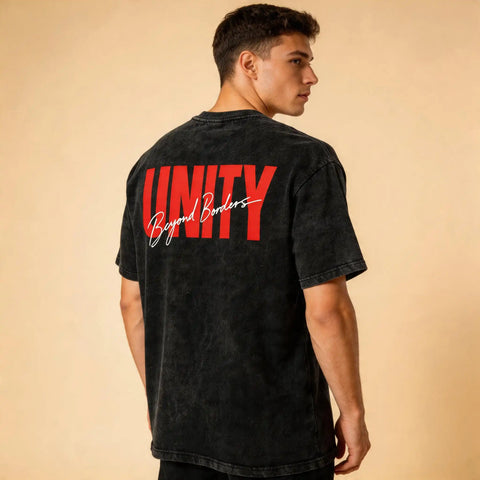 Back view of a male model wearing the Celvian Beyond Borders T-Shirt in washed black, featuring a bold red UNITY graphic with white Beyond Borders script on a clean beige studio background.