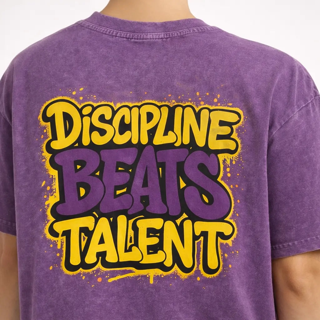 Close-up of back print on purple washed oversized t-shirt reading ‘Discipline Beats Talent’ in bold graffiti style.