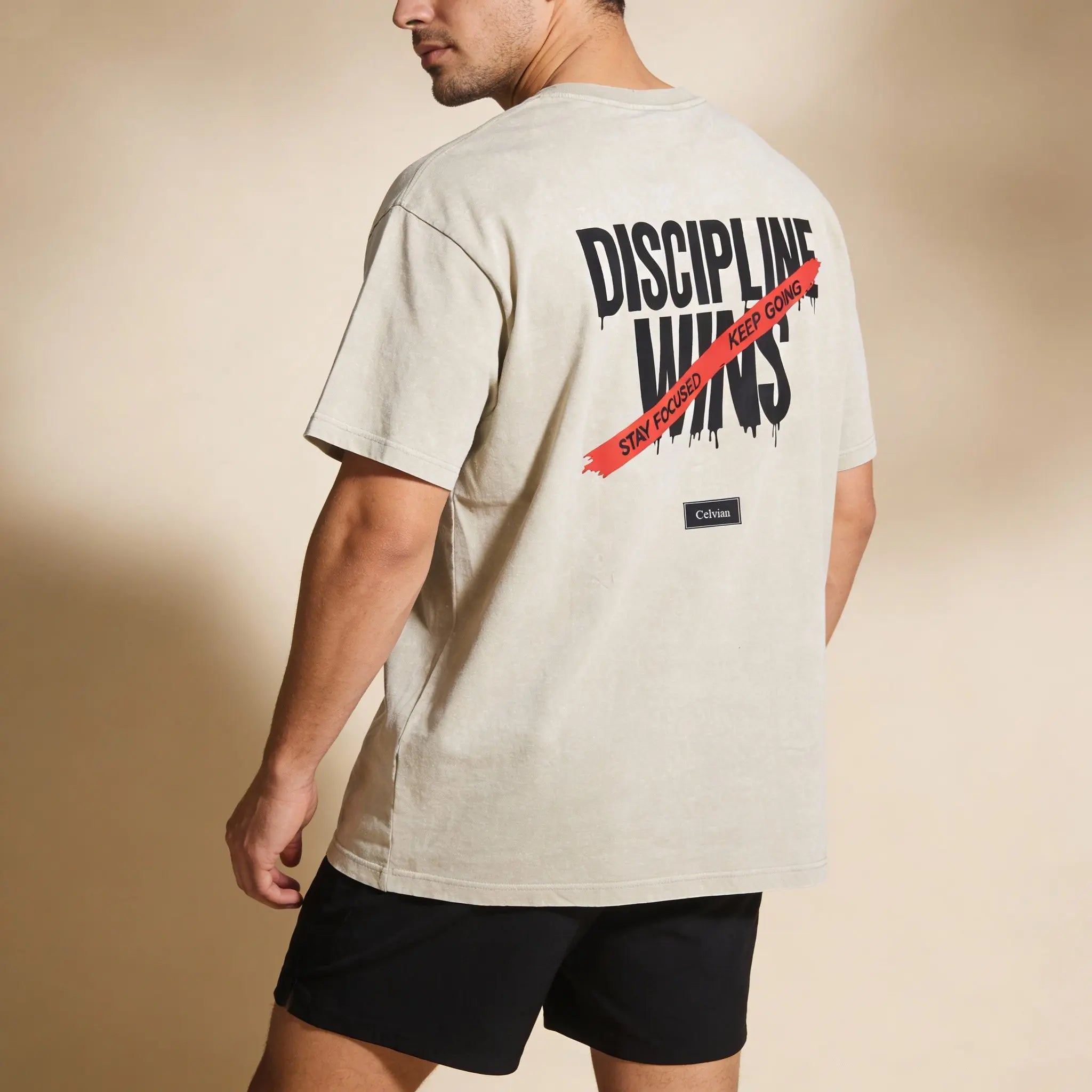 Angled back view of beige oversized t-shirt with ‘Discipline Wins’ print and red ‘Stay focused, keep going’ stripe, Celvian label.