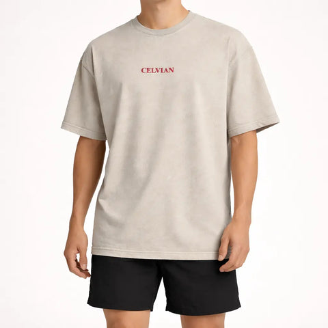 Front view of beige oversized t-shirt with small red Celvian logo on chest, worn with black shorts.