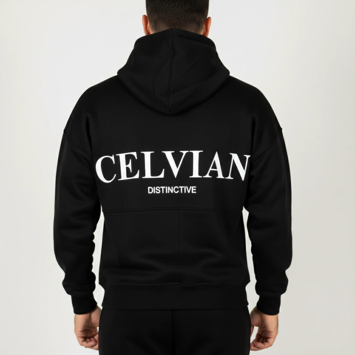 Celvian Distinctive black hoodie, back view showing large embroidered CELVIAN and DISTINCTIVE logos, minimalist studio setting.