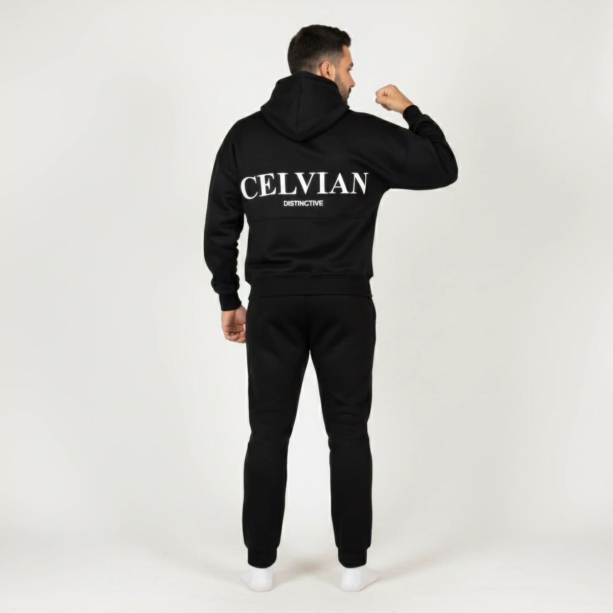 Celvian Distinctive black tracksuit, back view showing embroidered CELVIAN logo on hoodie, model flexing arm, minimalist studio setting.