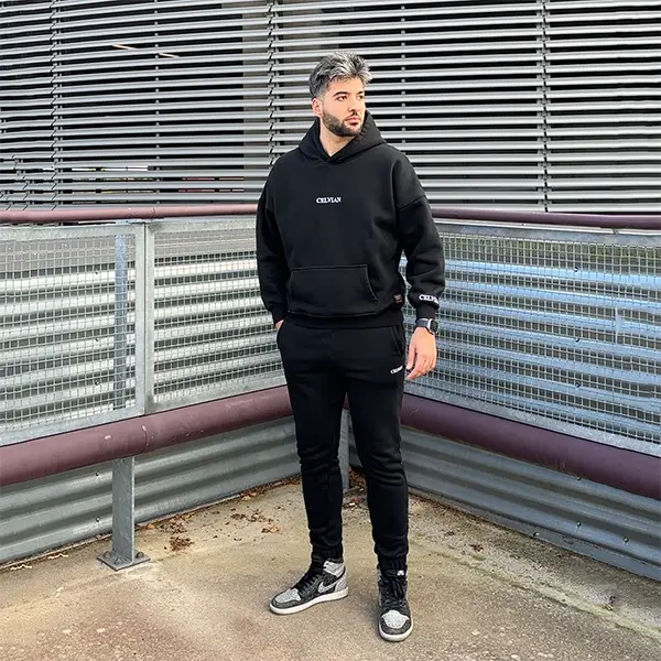 Male model wearing Celvian black tracksuit with oversized hoodie and slim joggers, CELVIAN logo on chest, sleeve and leg, standing on an industrial rooftop against metal railings, looking away from the camera.
