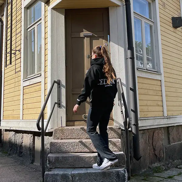Female model in Celvian black tracksuit, back view of oversized hoodie with large CELVIAN logo, walking up stone steps to a yellow wooden house, casual street style scene.