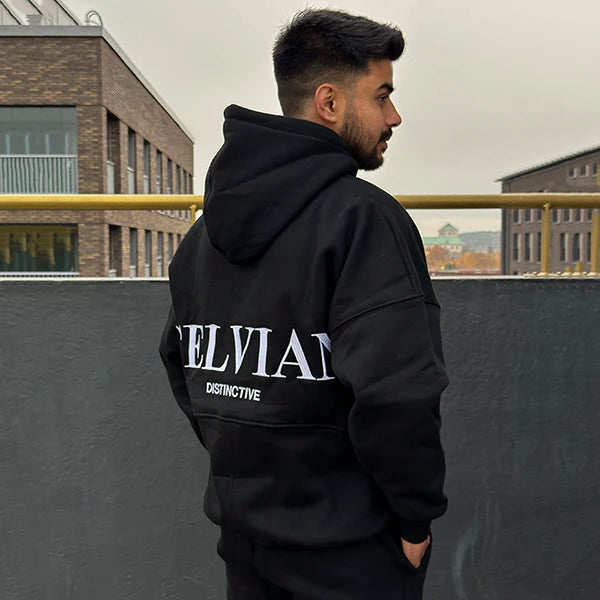 Male model wearing Celvian Distinctive black tracksuit, back view of oversized hoodie with large CELVIAN logo, standing on an urban rooftop.