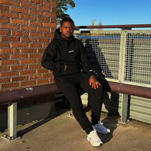 Male model wearing Celvian black tracksuit with oversized hoodie and joggers, sitting on an urban rooftop bench against a brick wall in direct sunlight.