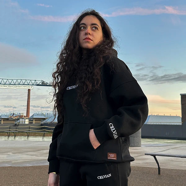 Female model wearing Celvian black tracksuit with oversized hoodie and pants, CELVIAN logo on chest, sleeve and leg, standing on urban rooftop at sunset looking away from the camera.