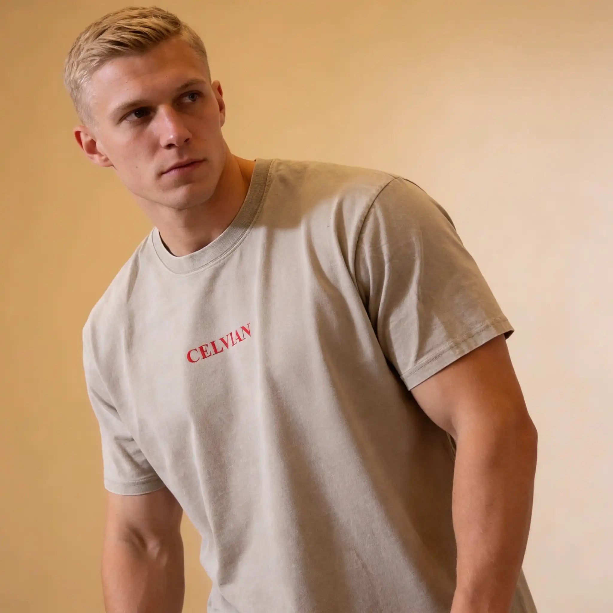 Male model wearing the Celvian Sandstone Global Vision T-Shirt in a washed beige finish, shown from a front angled view with the minimal red CELVIAN chest logo.