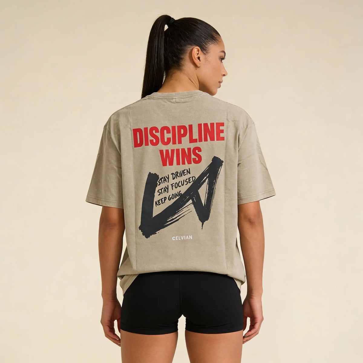 Back view of a woman wearing the Celvian Stay Driven T-Shirt in washed beige, featuring the bold ‘Discipline Wins’ graphic print, oversized fit, and black shorts.