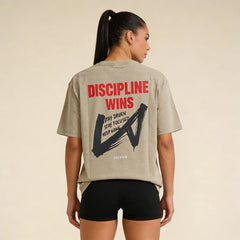 Back view of a woman wearing the Celvian Stay Driven T-Shirt in washed beige, featuring the bold ‘Discipline Wins’ graphic print, oversized fit, and black shorts.