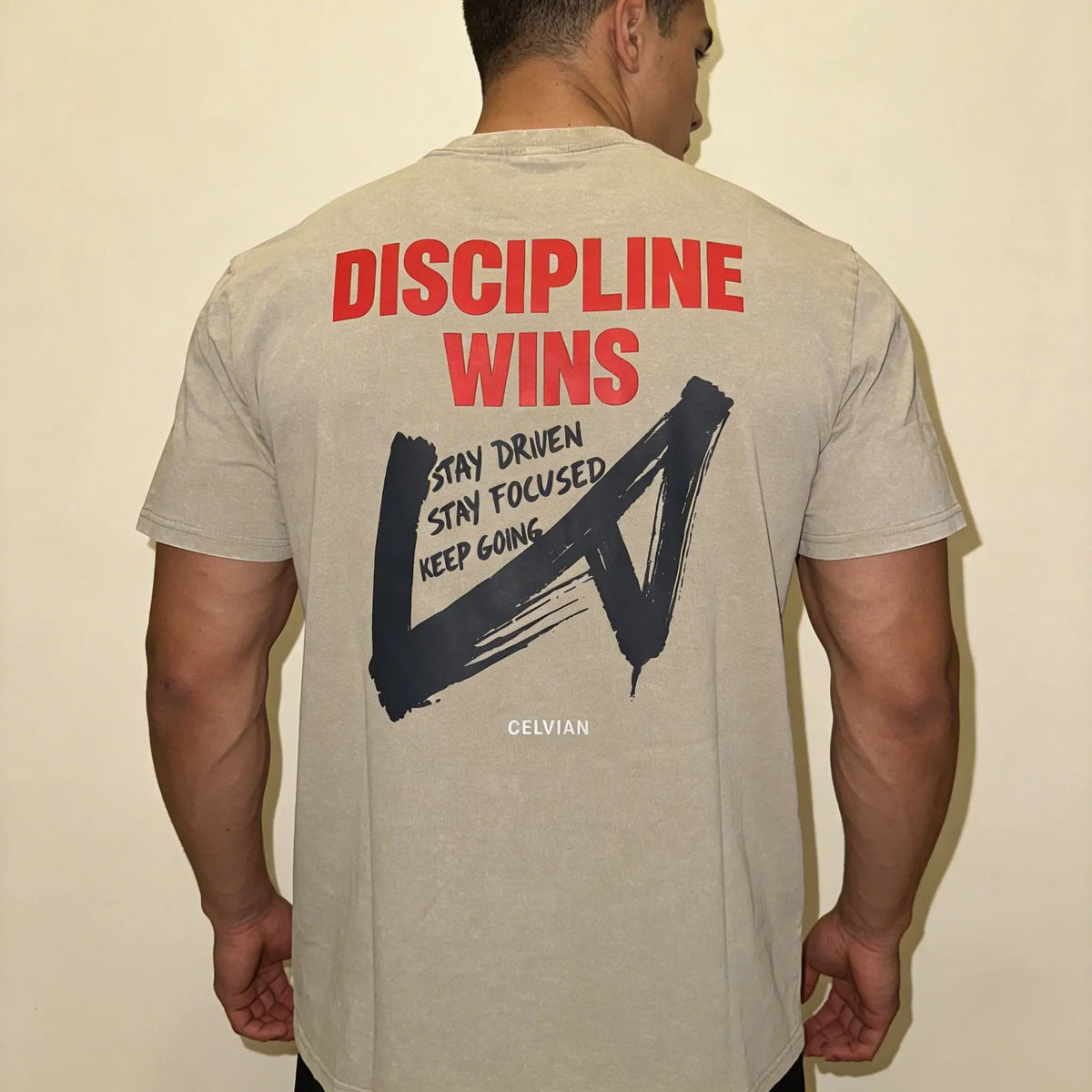 Back view of a male model wearing the Celvian Stay Driven T-Shirt in sandstone beige, featuring a bold red ‘DISCIPLINE WINS’ graphic, dark brushstroke design, and motivational back print.